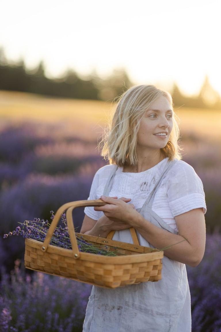 In a lavender field at golden hour, a woman holds a woven basket of freshly cut lavender.